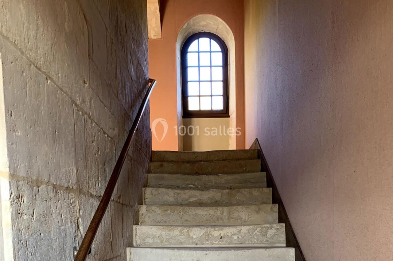 Escalier en pierre avec une rampe en métal, menant à une fenêtre cintrée laissant passer la lumière naturelle.