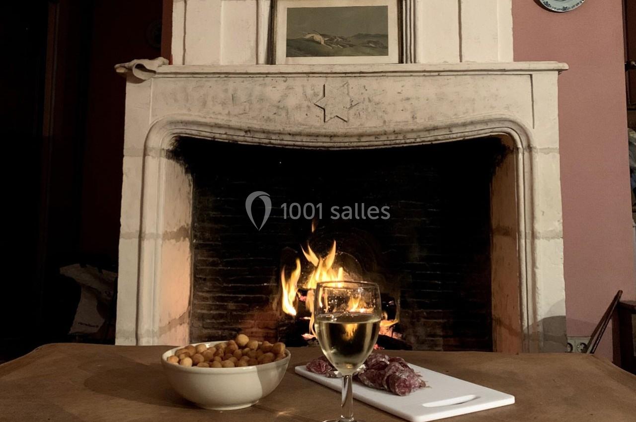 Verre de vin blanc, planche de charcuterie et bol d'amuse-bouches posés sur une table devant une cheminée allumée.