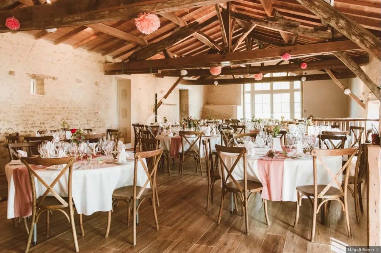 Salle de réception rustique avec tables rondes décorées de nappes blanches et chaises en bois sous une charpente apparente.