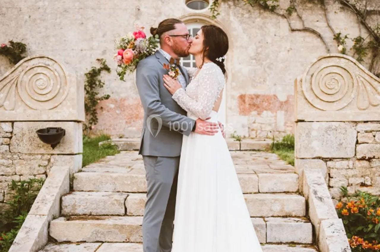 Un couple en tenue de mariage s'embrasse sur des marches en pierre devant un mur ancien orné de végétation.