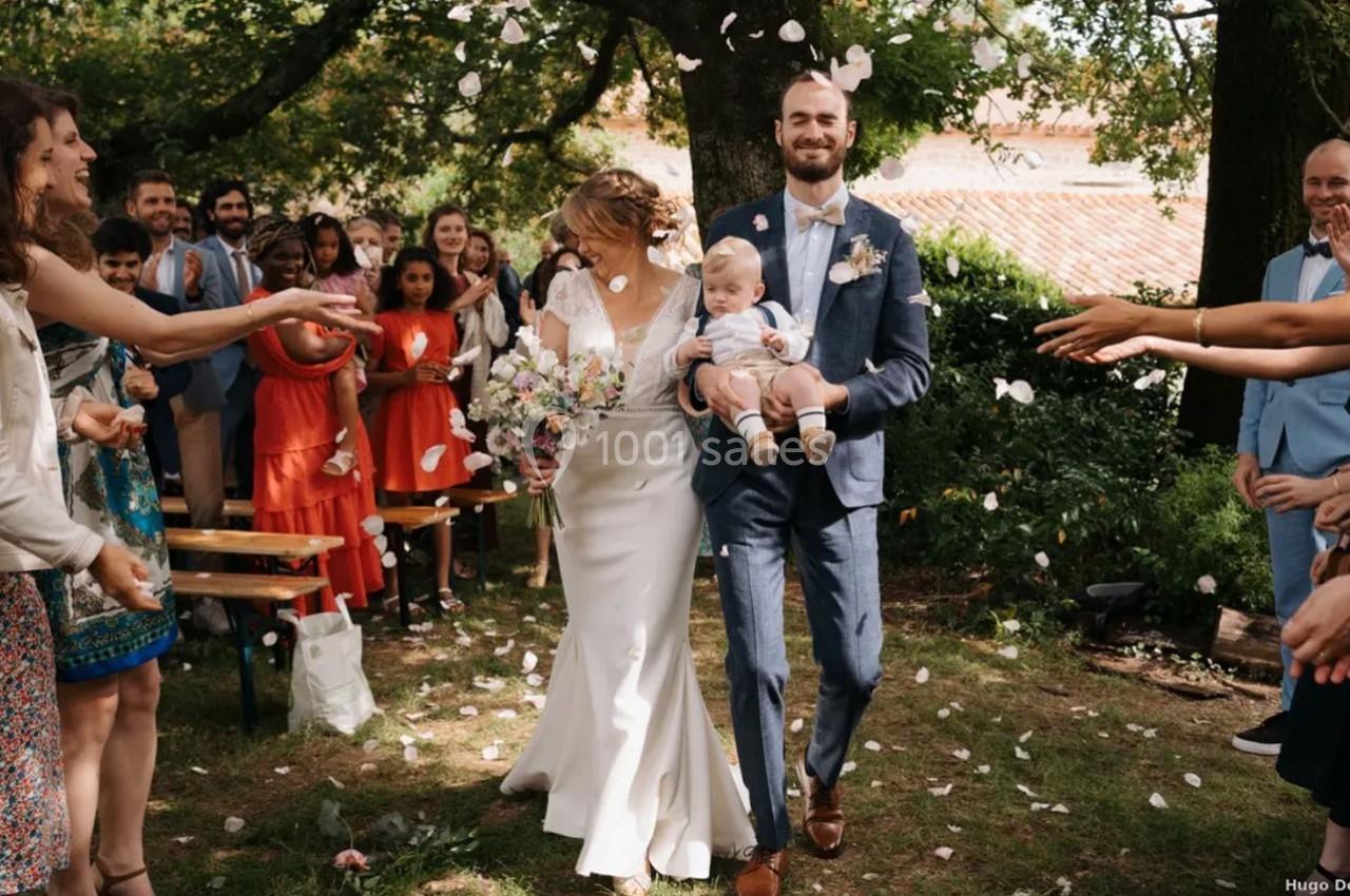 Un couple marié souriant marche sous une pluie de pétales, entouré d'invités dans un jardin.