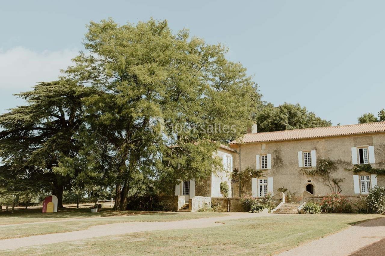 Grande maison en pierre avec volets blancs, entourée d'arbres et d'un jardin sous un ciel dégagé.