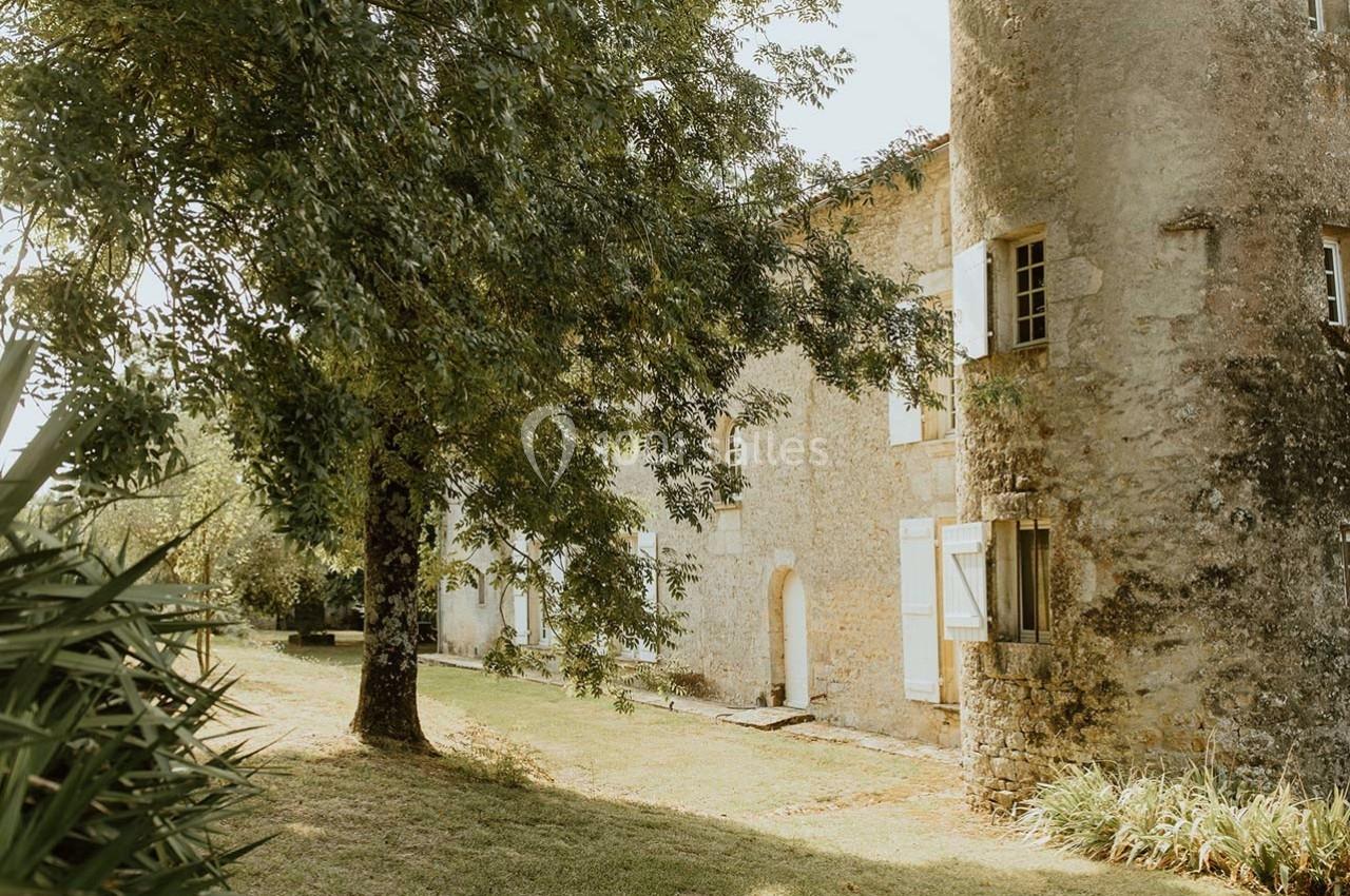 Façade en pierre d'une maison ancienne avec volets blancs, entourée d'arbres et d'un jardin ensoleillé.