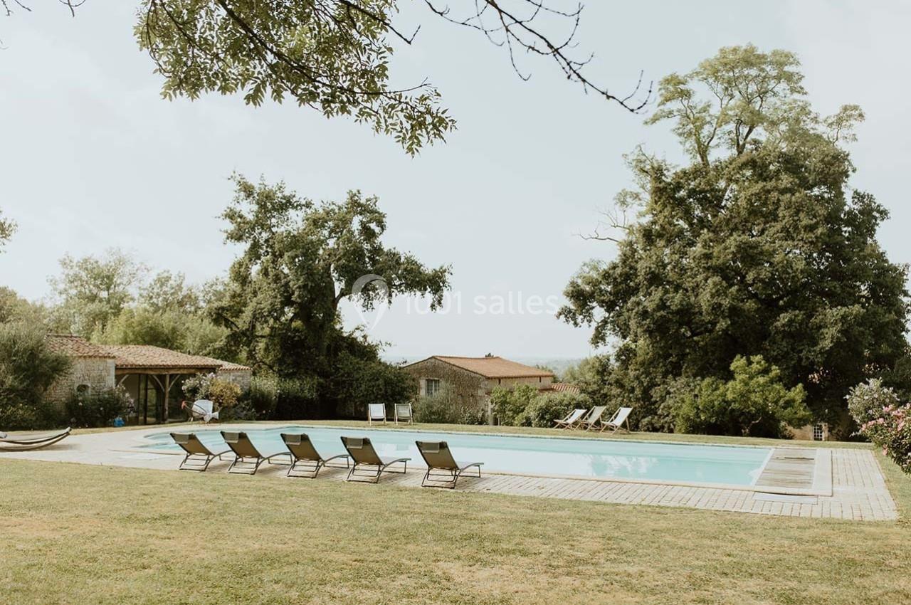Piscine extérieure entourée de chaises longues, pelouse et arbres, avec des bâtiments en pierre en arrière-plan.