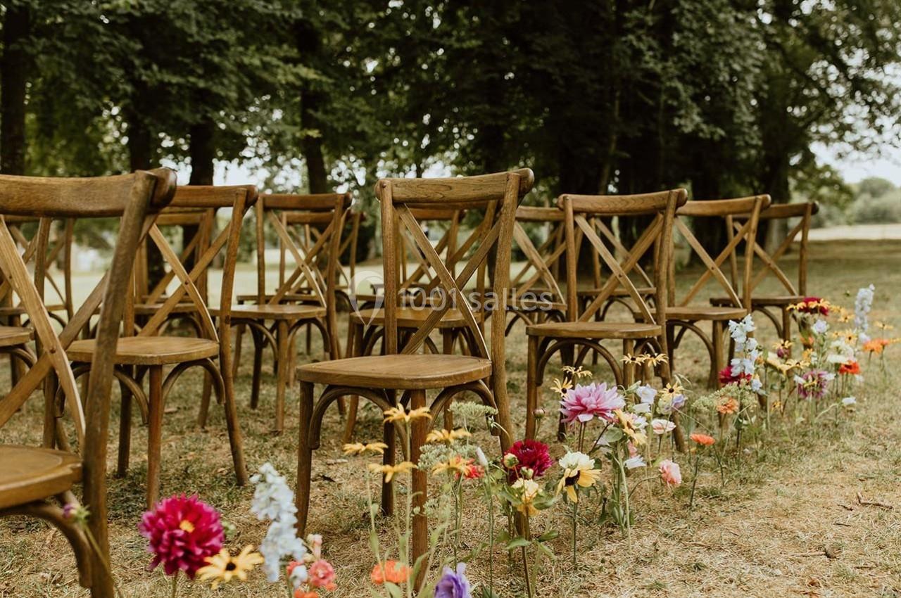 Chaises en bois disposées en rangées sur une pelouse, entourées de fleurs colorées, près d'arbres.