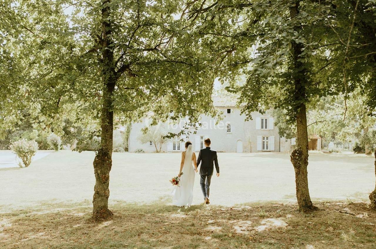 Un couple marche main dans la main sur une pelouse verdoyante, entouré d'arbres, avec un bâtiment en pierre en arrière-plan.