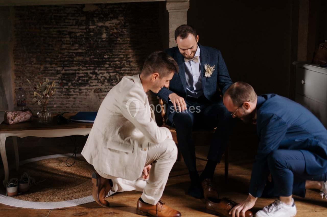 Deux hommes ajustent les chaussures d'un troisième homme assis, dans une pièce au décor rustique.