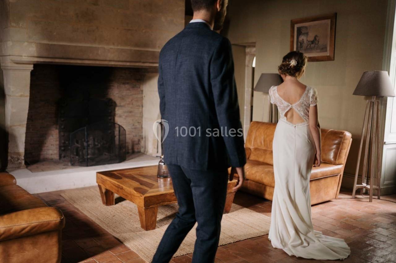 Un homme en costume marche vers une femme en robe de mariée dans un salon avec cheminée et meubles en cuir.