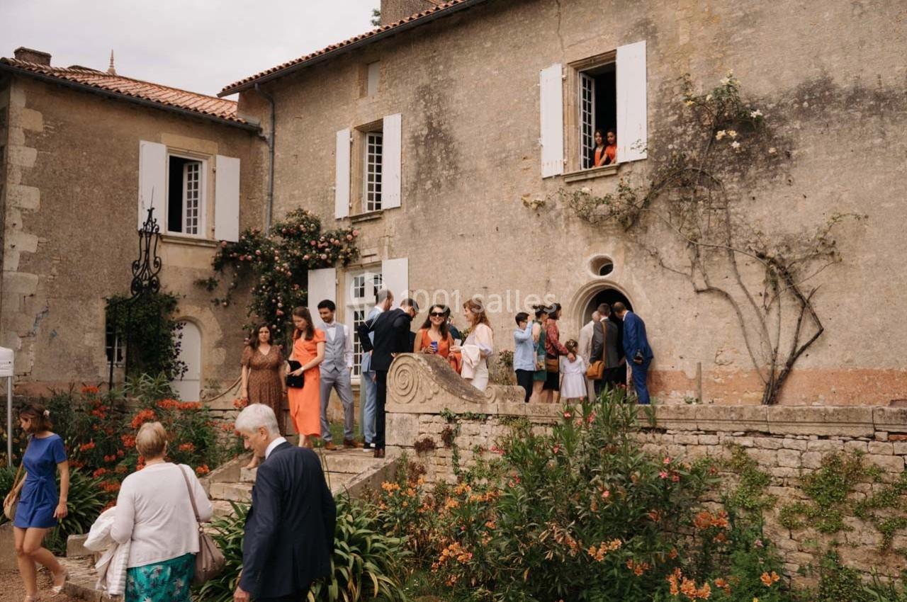Un groupe de personnes discute devant une maison ancienne avec des murs en pierre et des fleurs grimpantes.