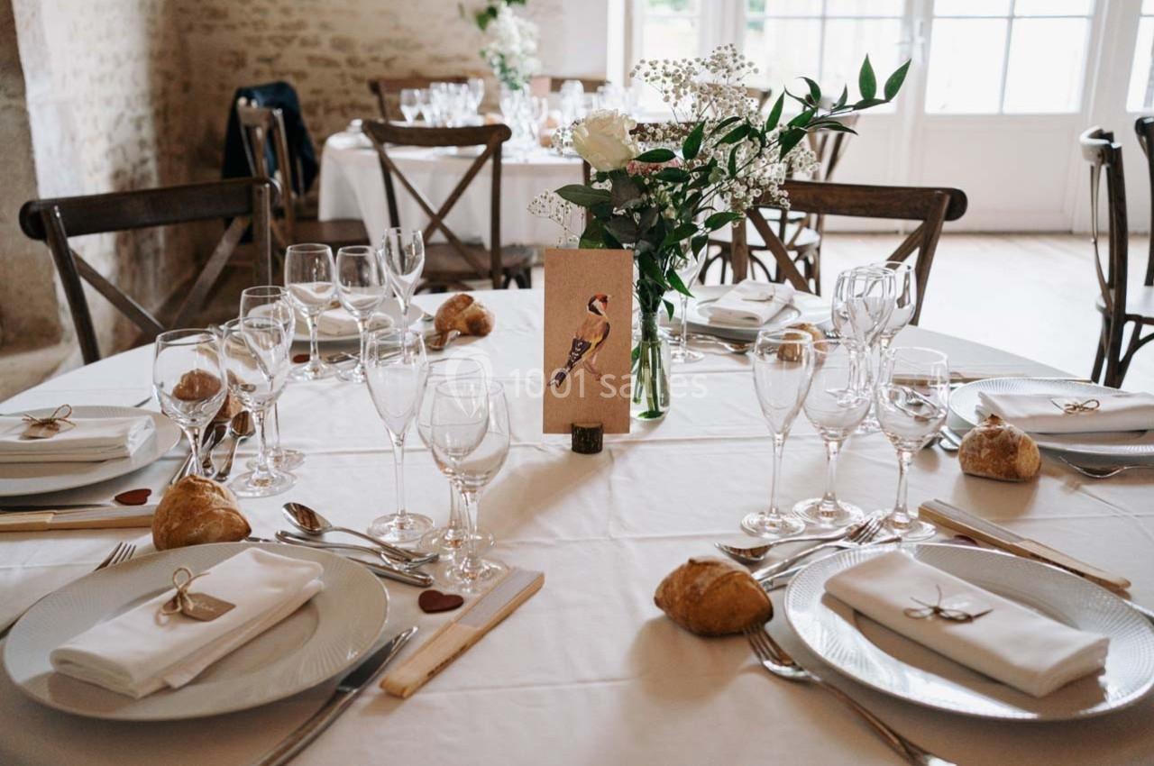 Table dressée avec nappes blanches, vaisselle élégante, verres, fleurs et marque-place décoré d'un oiseau.