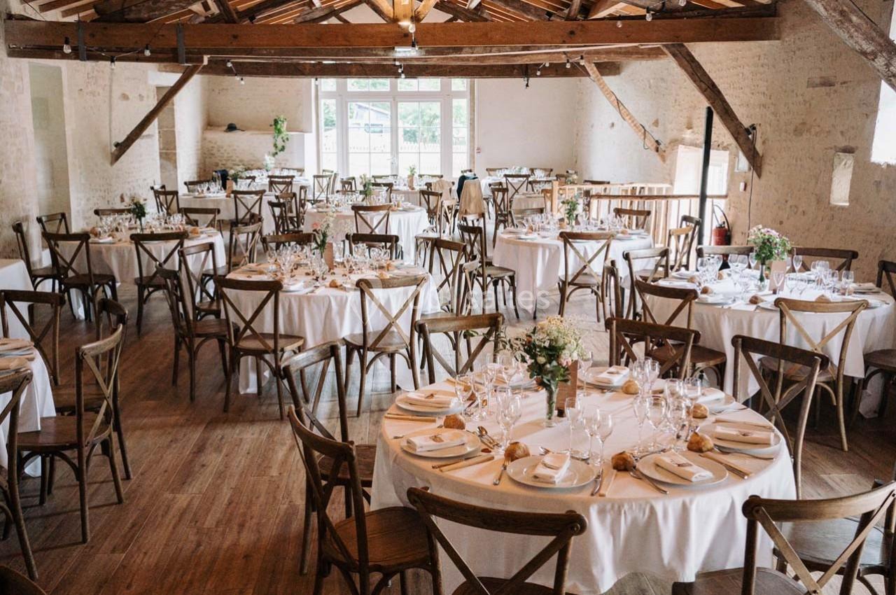 Salle de réception lumineuse avec tables rondes dressées, chaises en bois et poutres apparentes.