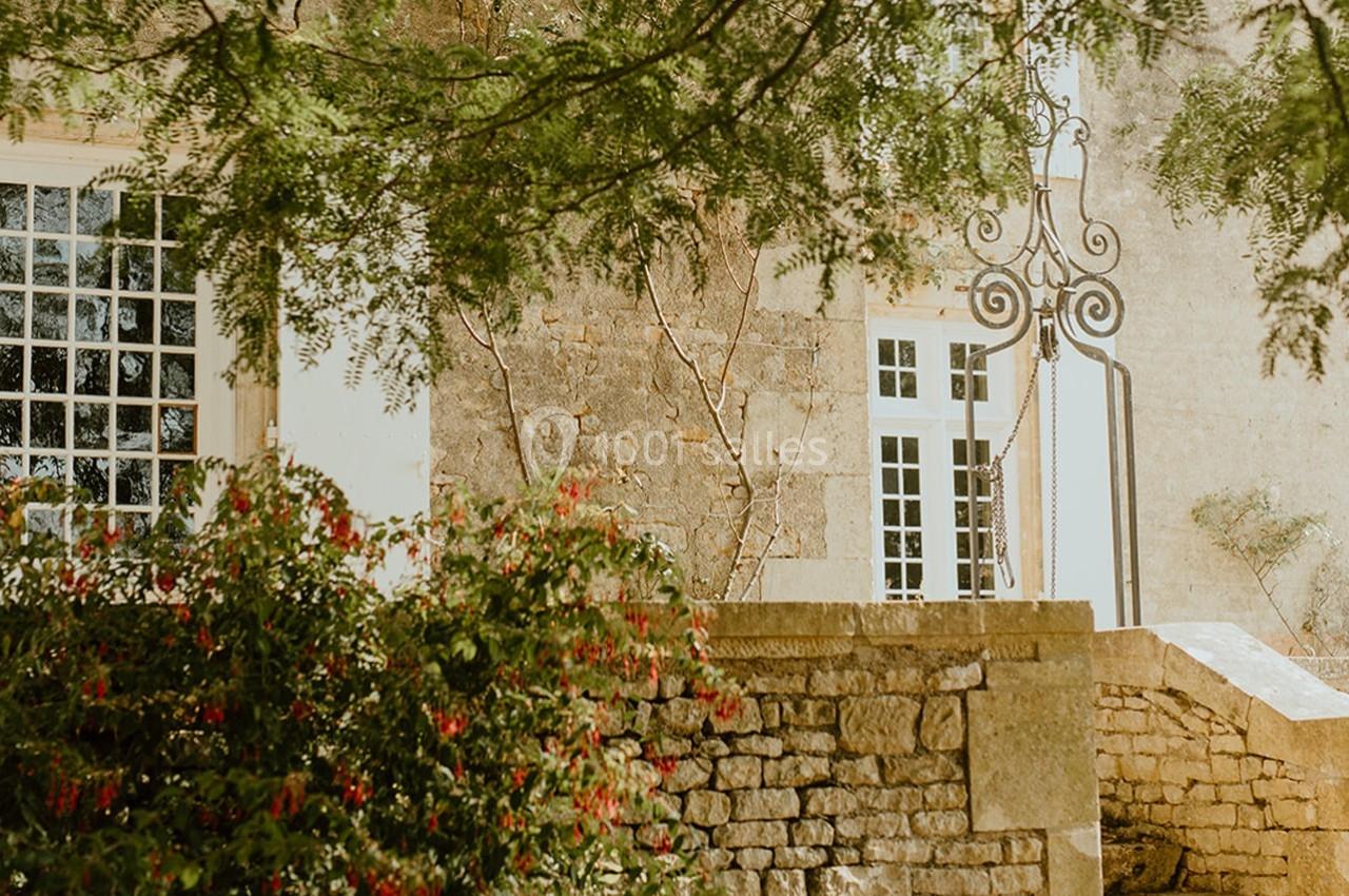 Façade en pierre d'un bâtiment ancien avec fenêtres blanches, entourée de verdure et d'un mur en pierre.