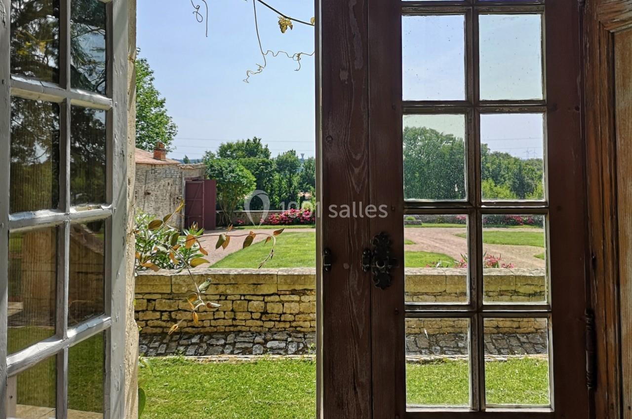 Vue depuis une porte-fenêtre ouverte sur un jardin verdoyant avec pelouse, buissons fleuris et muret en pierre.