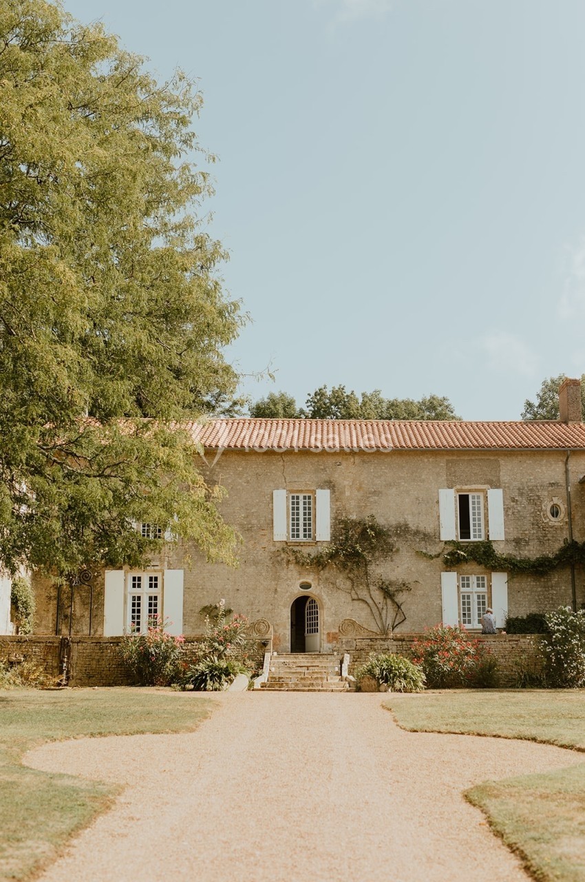 Façade d'une maison en pierre avec volets blancs, entourée de végétation et d'un chemin gravillonné.