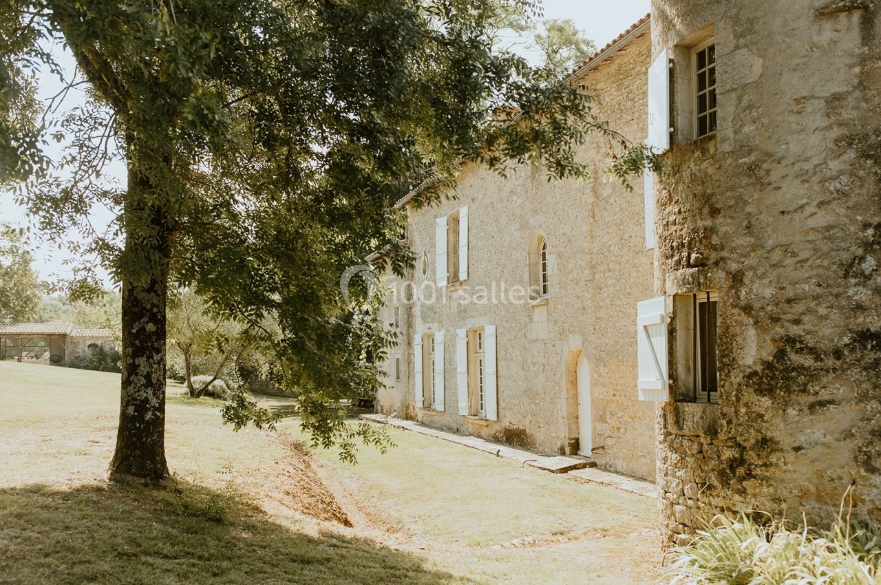 Façade en pierre d'une maison ancienne entourée d'arbres et d'une pelouse sous une lumière douce.