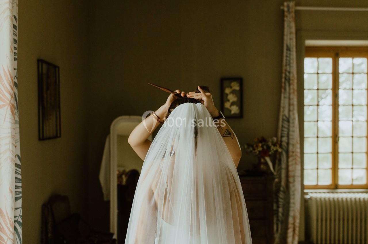 Une mariée de dos ajuste son voile dans une chambre lumineuse avec des rideaux et un miroir en arrière-plan.