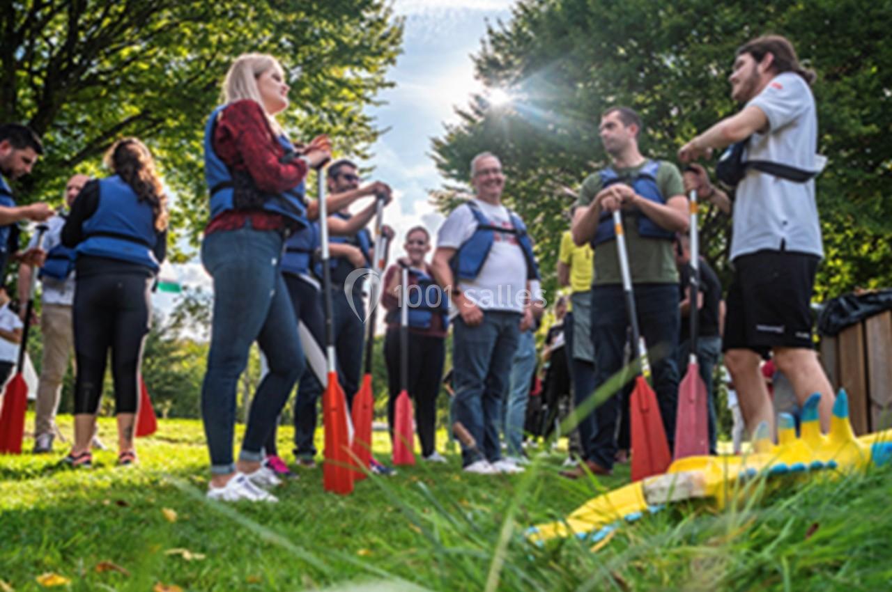 Un groupe de personnes en gilets de sécurité discute sur une pelouse, tenant des pagaies, sous un ciel ensoleillé.