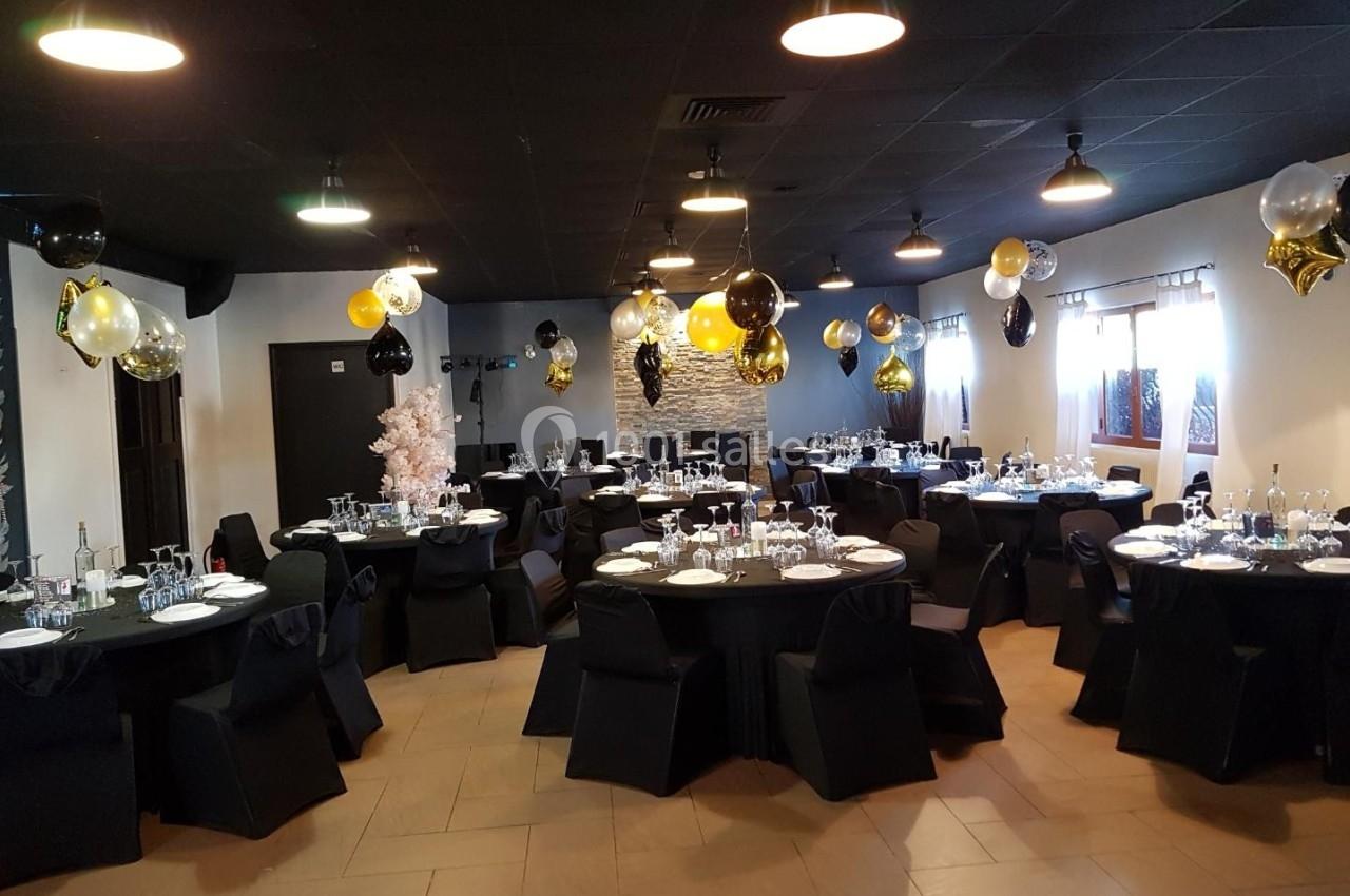 Salle décorée pour un événement avec des tables rondes dressées, des nappes noires et des ballons dorés, noirs et blancs.
