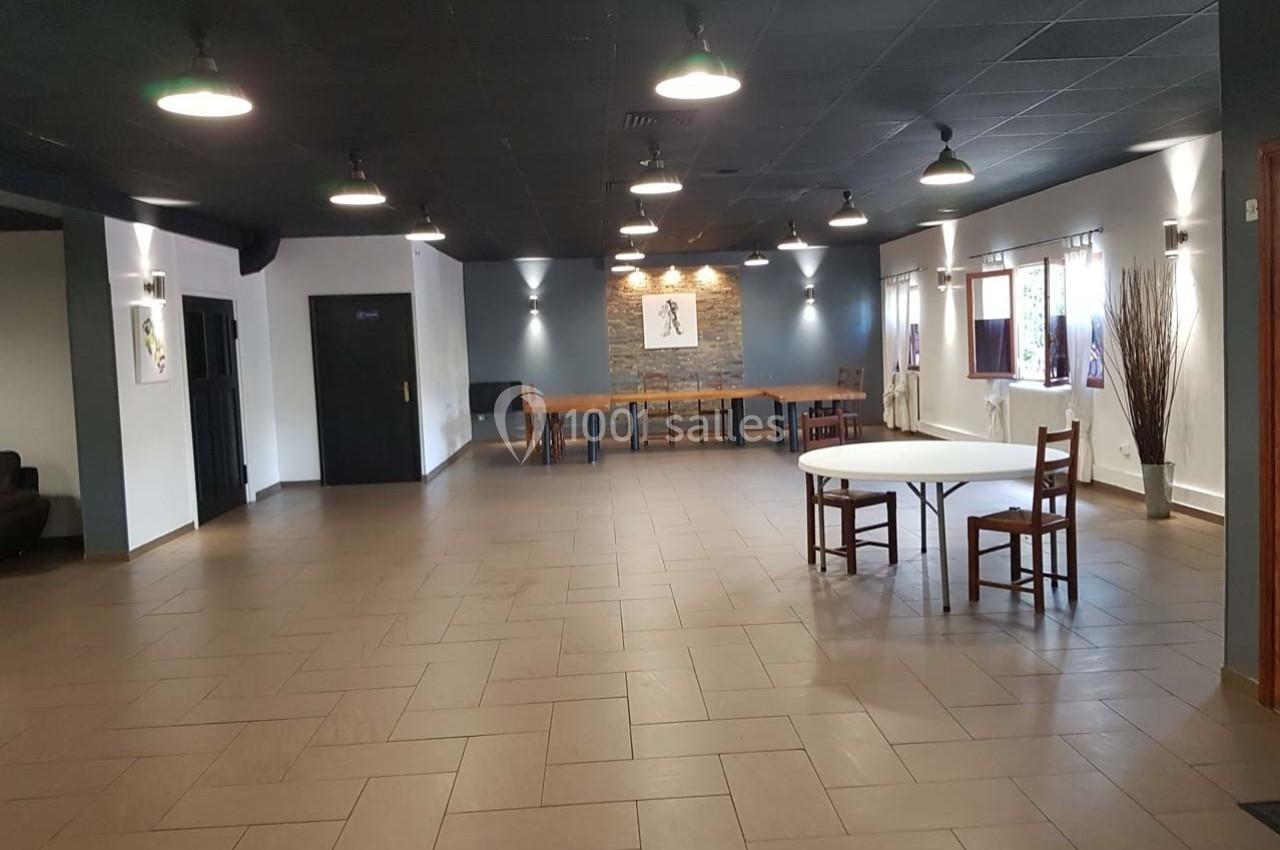 Salle spacieuse avec sol carrelé, plafond noir, tables et chaises dispersées, murs clairs et éclairage suspendu.