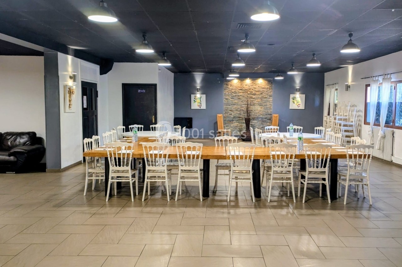 Salle spacieuse avec tables en bois alignées, chaises blanches, murs clairs et éclairage suspendu.