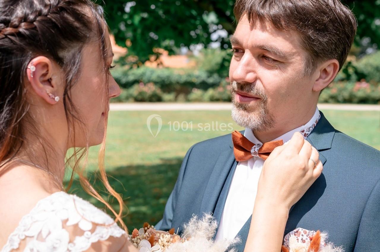 Une femme ajuste le nœud papillon d'un homme en costume lors d'une cérémonie en extérieur.