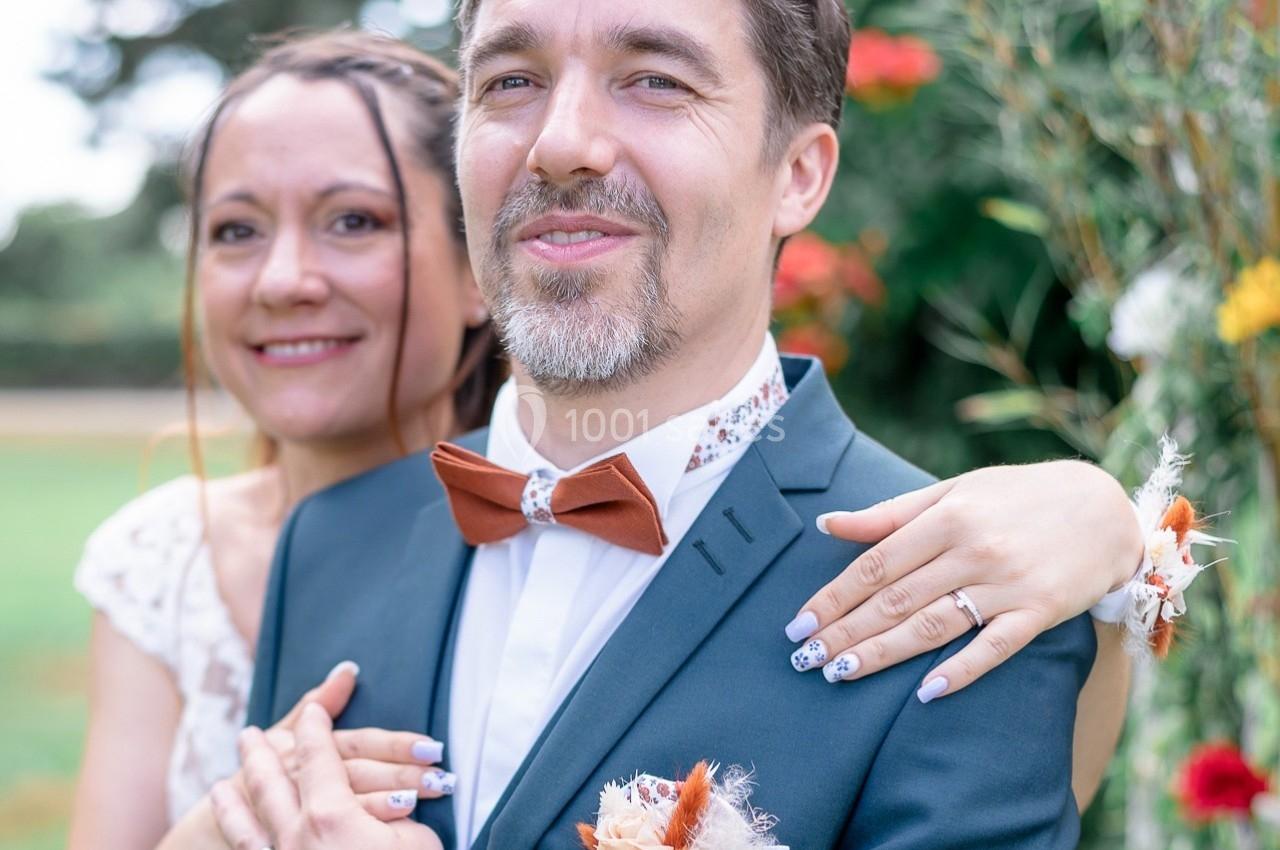 Un couple souriant en tenue de mariage pose devant un décor extérieur fleuri.
