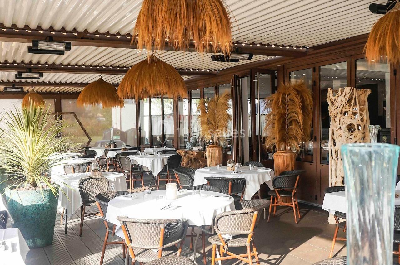 Terrasse d'un restaurant avec tables dressées, chaises en rotin, luminaires suspendus et décoration végétale.