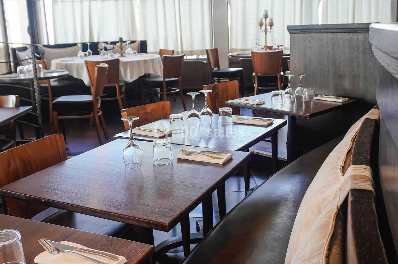 Salle de restaurant lumineuse avec tables en bois dressées, chaises et banquettes, grandes fenêtres en arrière-plan.