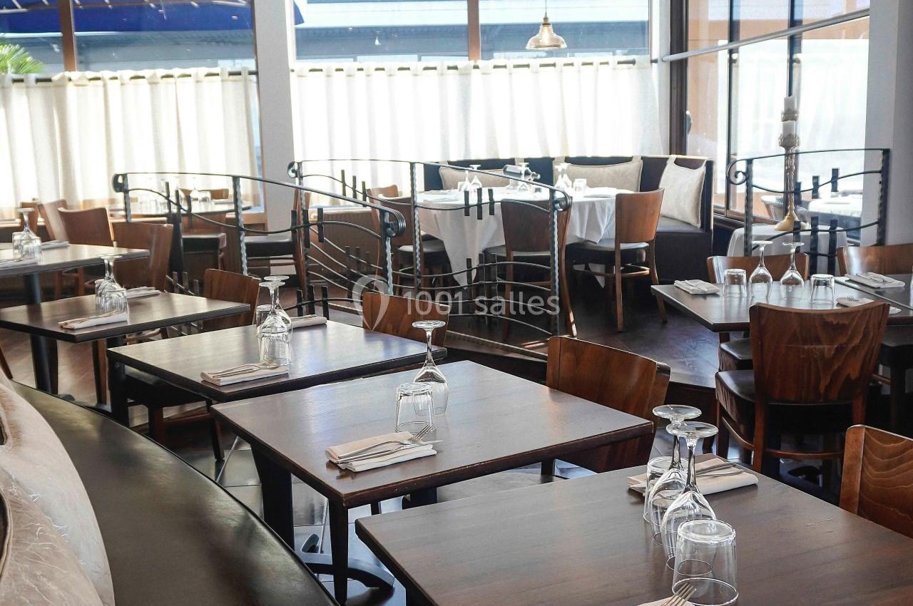 Salle de restaurant lumineuse avec tables en bois dressées, chaises assorties et grandes baies vitrées.