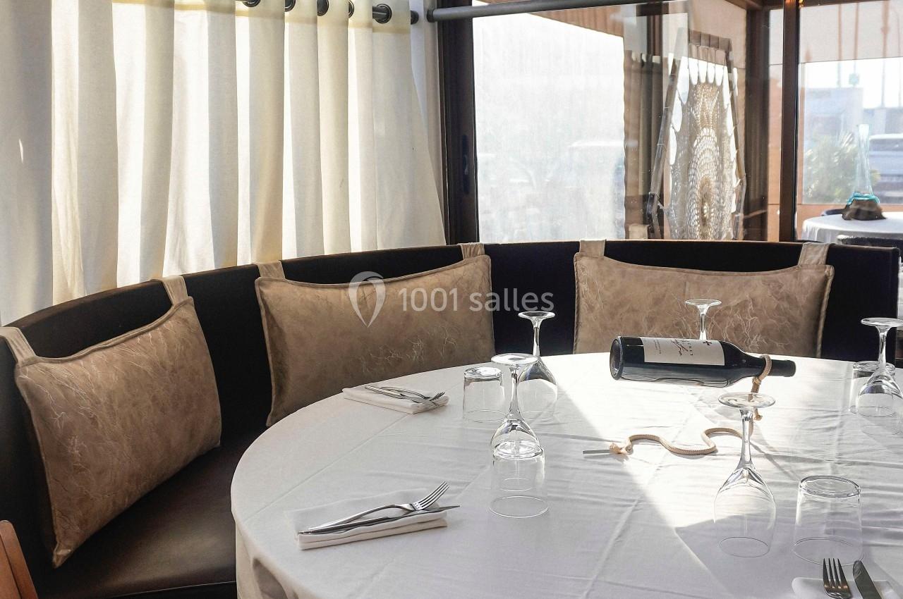 Table ronde dressée avec nappe blanche, verres et couverts, entourée de banquettes et éclairée par une lumière naturelle.