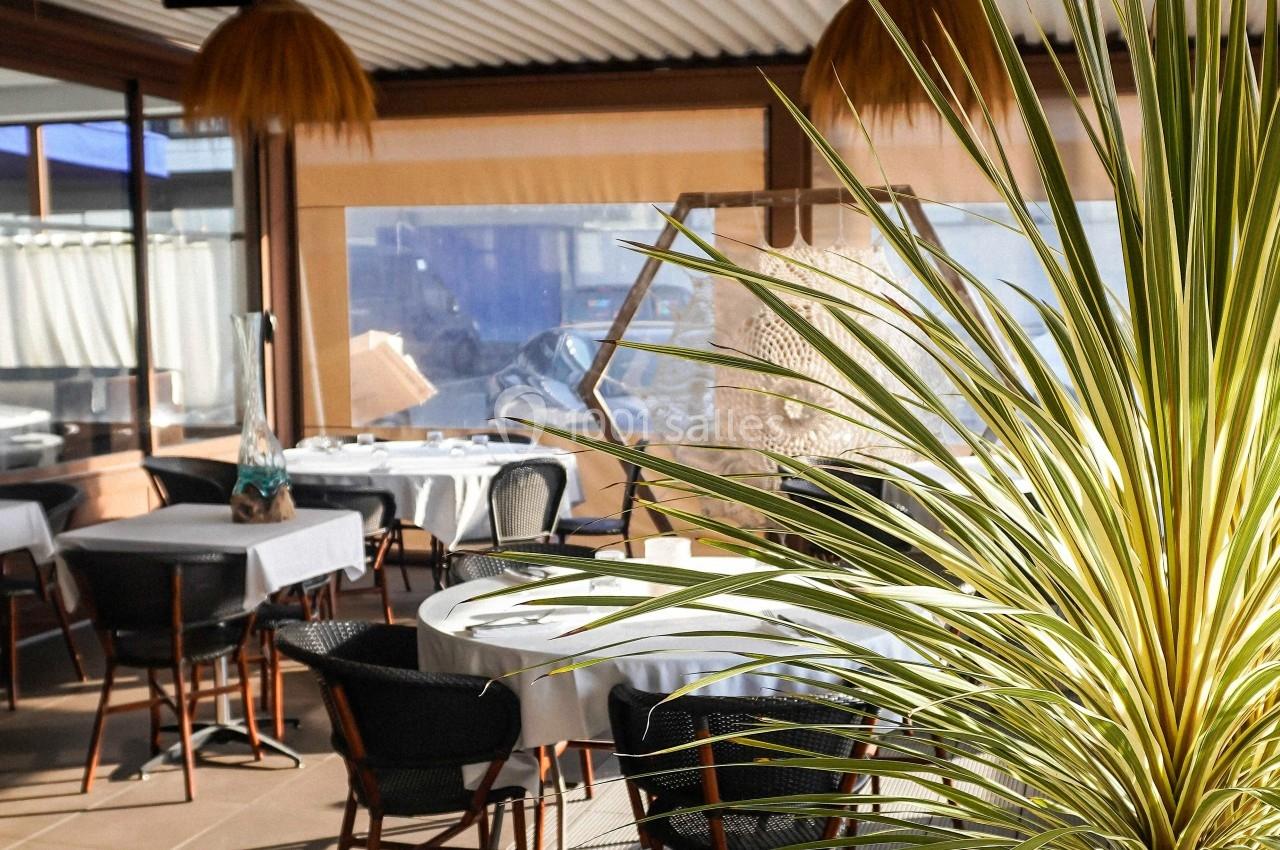 Salle lumineuse avec des tables rondes dressées, des chaises en osier et une plante verte au premier plan.