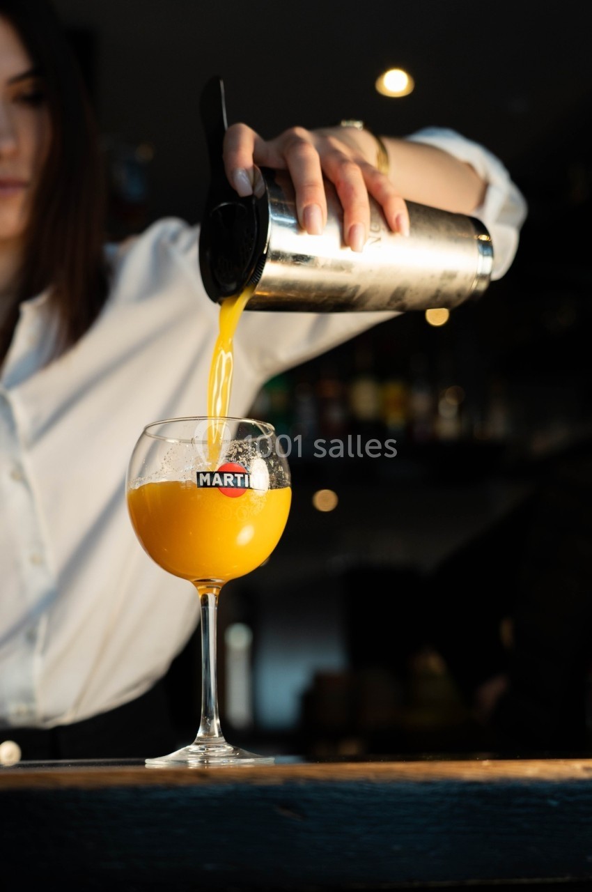 Une personne verse un cocktail jaune vif dans un verre à pied dans un environnement de bar sombre.