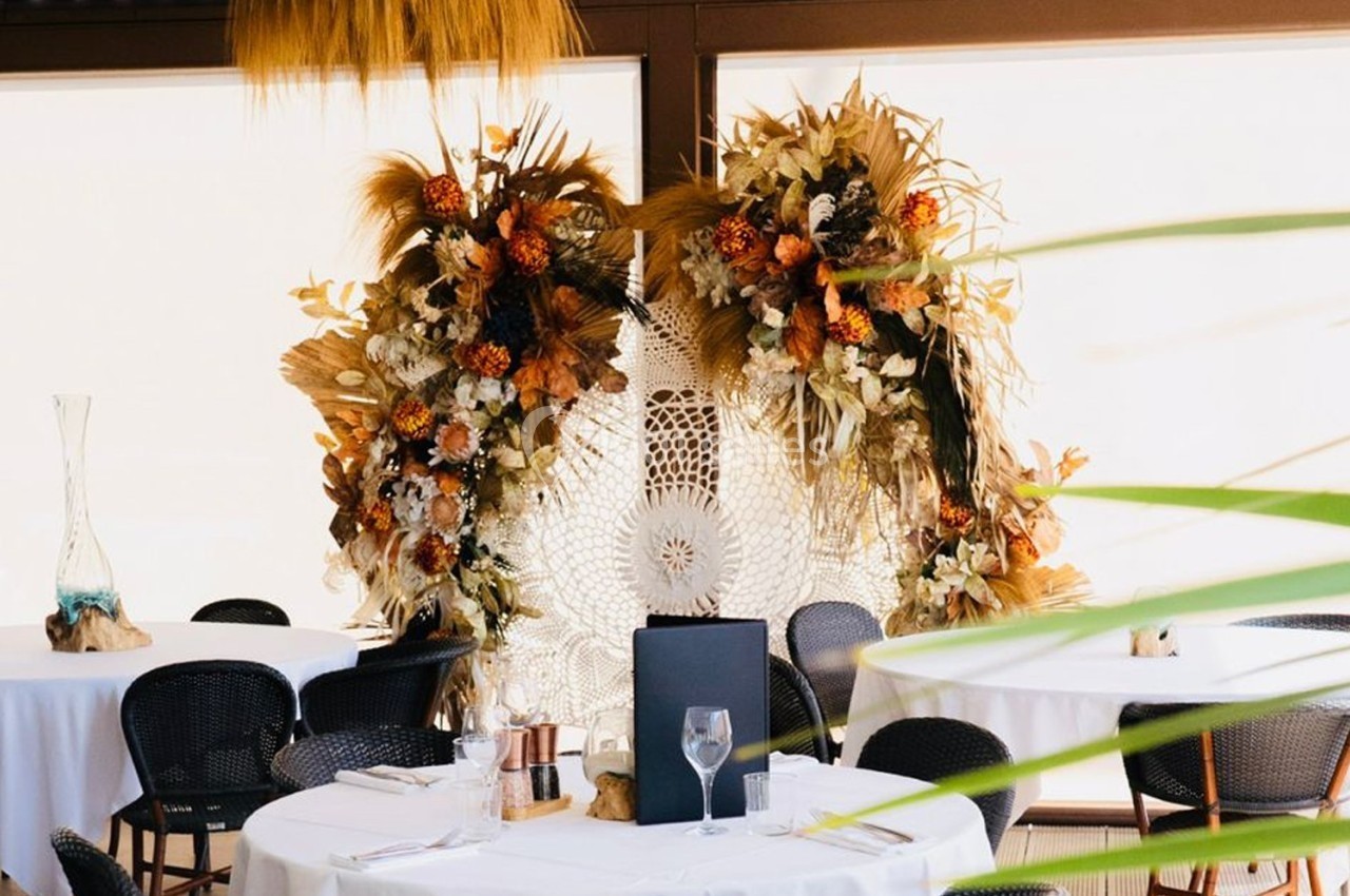 Décoration florale bohème avec des fleurs séchées et un macramé en arrière-plan, entourée de tables dressées.