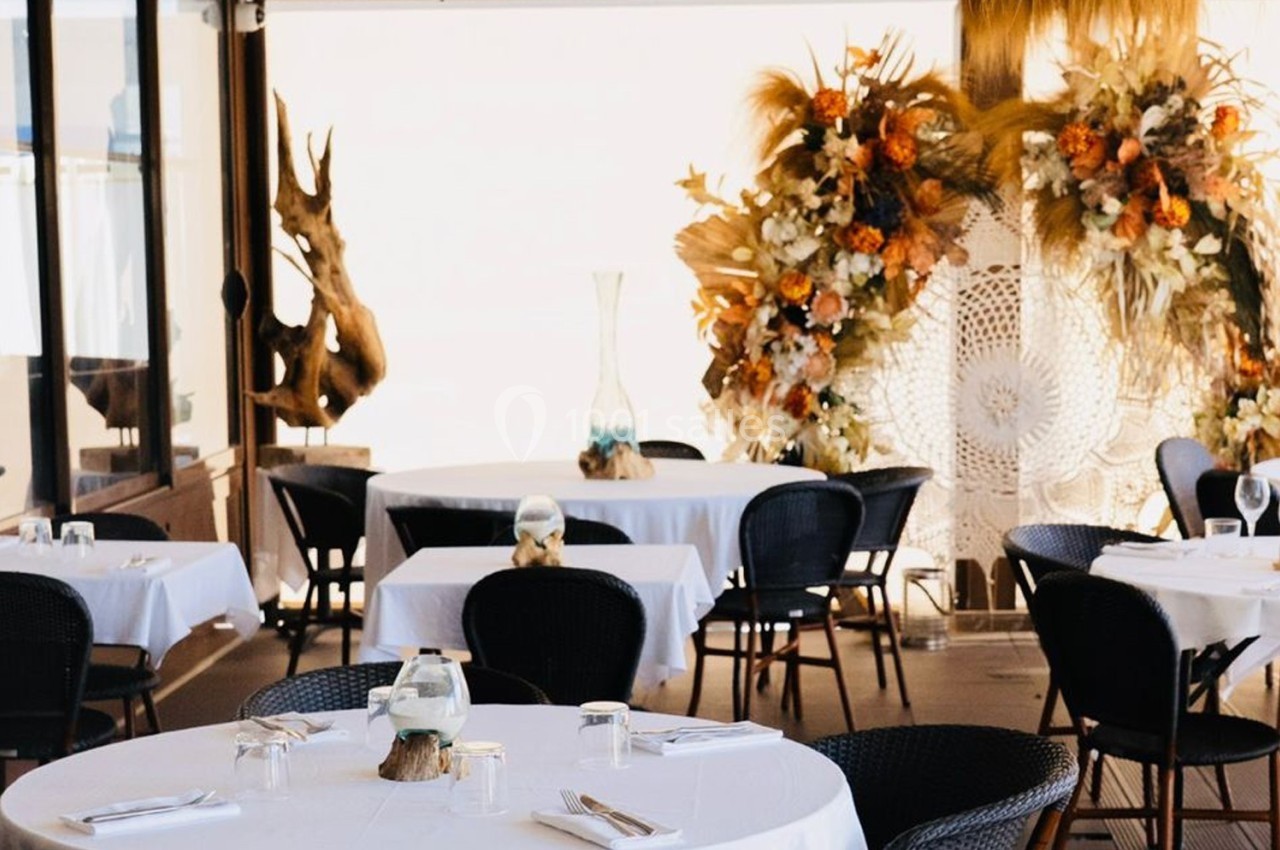 Salle de restaurant lumineuse avec tables dressées, chaises noires et décor floral aux tons chauds en arrière-plan.