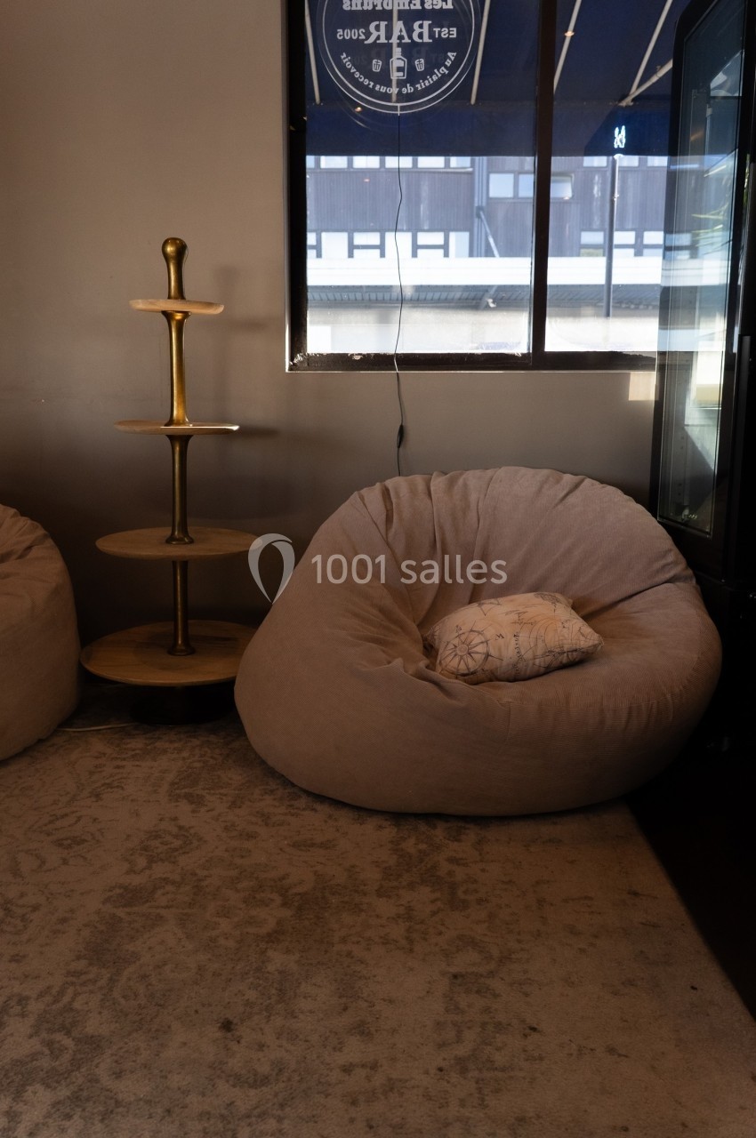 Fauteuil beige en forme de coussin avec un coussin décoratif, près d'une étagère en bois et d'une fenêtre.