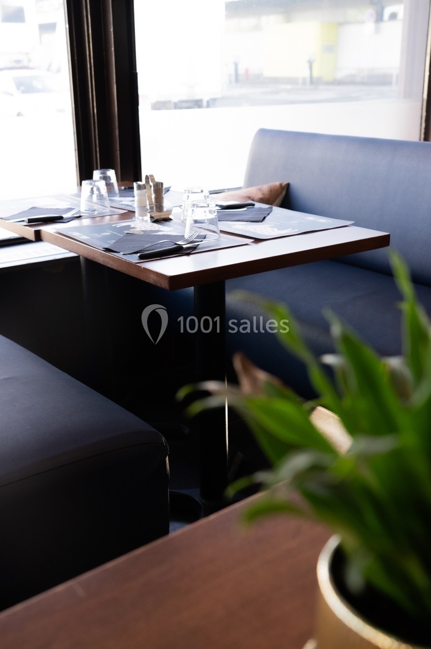 Table de restaurant dressée avec des verres et des serviettes, entourée de banquettes, près d'une fenêtre lumineuse.