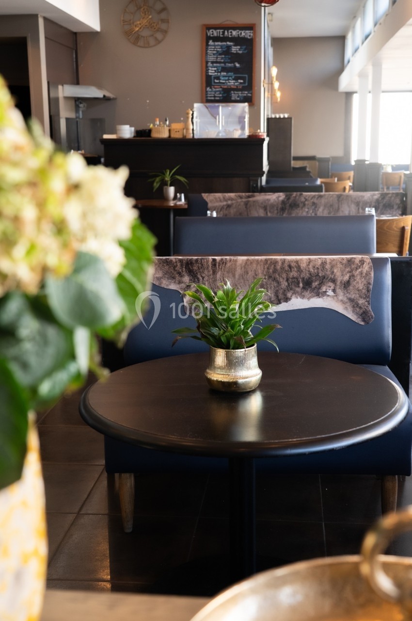 Table ronde avec une plante en pot au centre, située dans un restaurant lumineux avec des banquettes en arrière-plan.