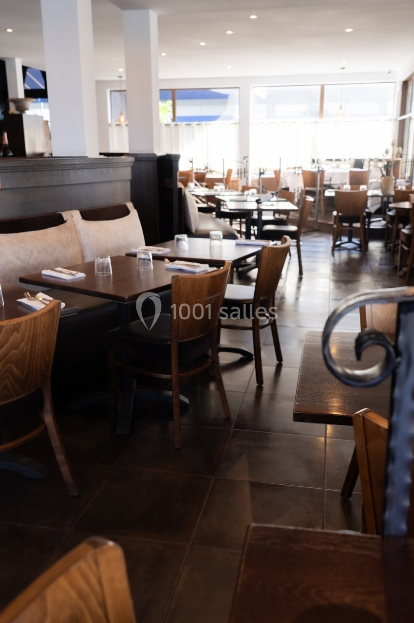 Salle de restaurant lumineuse avec tables en bois, chaises assorties et grandes baies vitrées en arrière-plan.