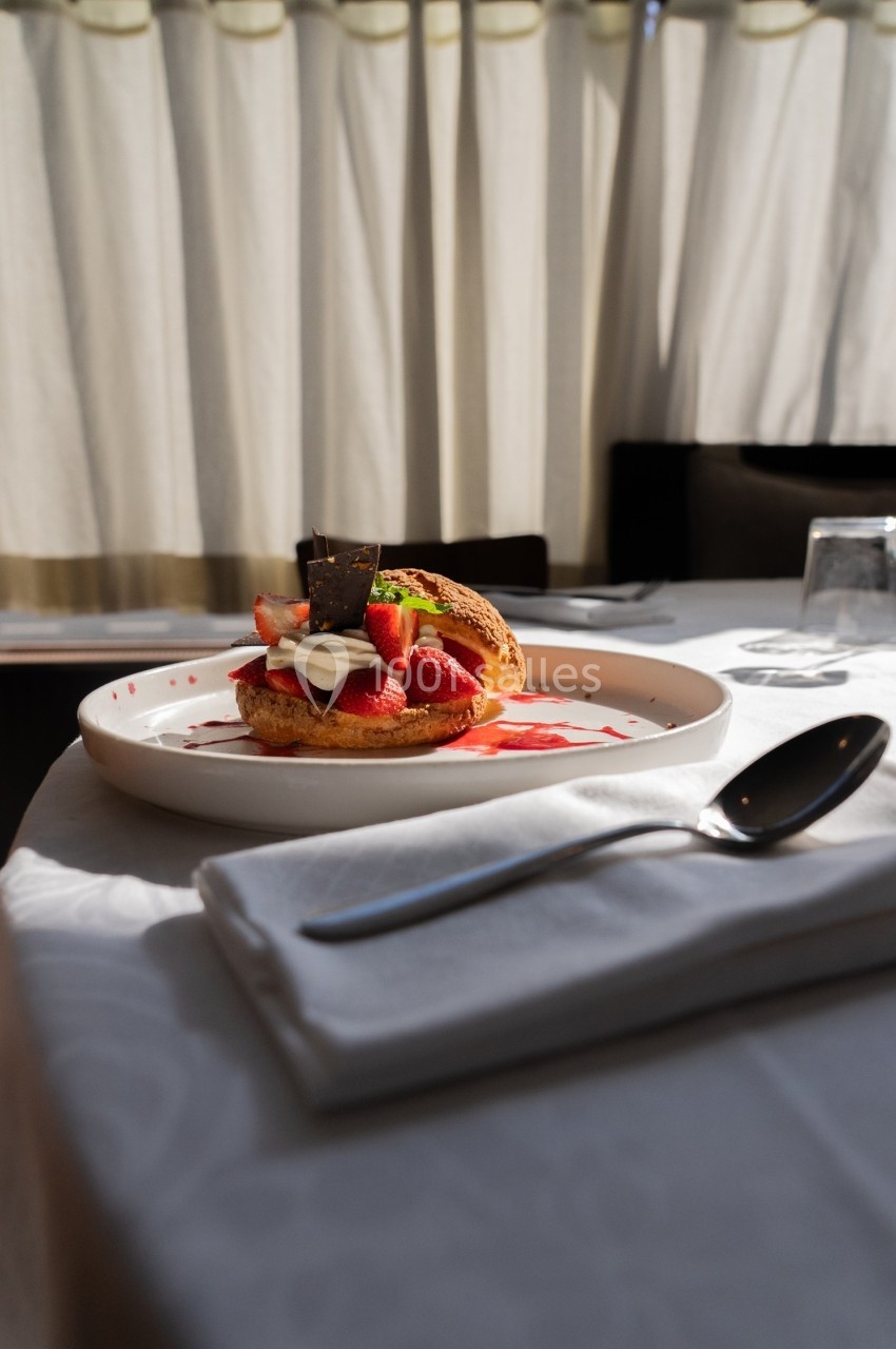 Dessert composé de fraises, crème et chocolat sur une assiette blanche, posé sur une table dressée près d'une fenêtre.