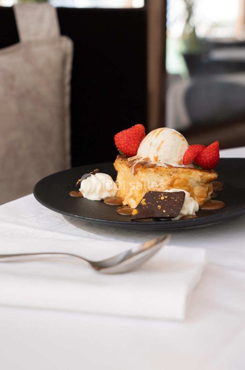 Dessert composé de brioche, glace vanille, fraises, chantilly, caramel et éclats de chocolat, servi sur une assiette noire.