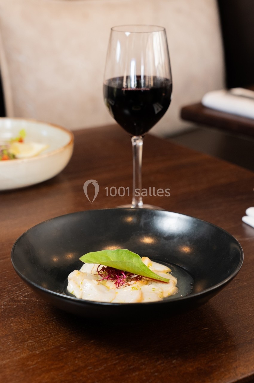 Assiette de carpaccio de poisson avec garniture verte, servie sur une table en bois avec un verre de vin rouge.