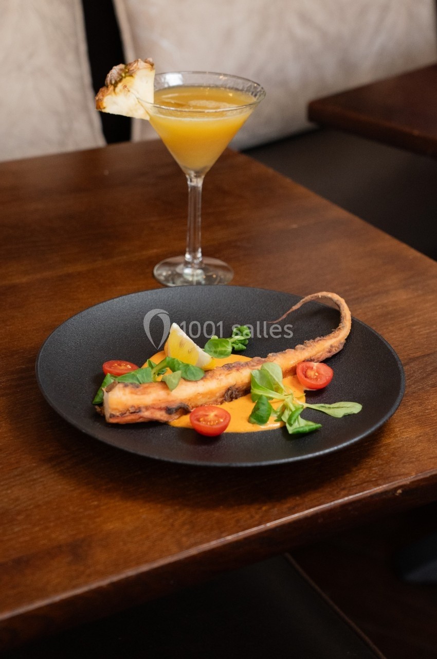 Assiette de tentacule de poulpe grillé avec sauce, tomates cerises et herbes, accompagnée d'un cocktail jaune.