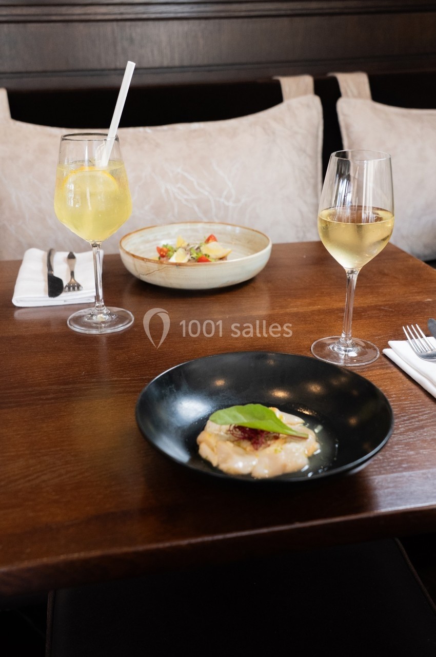 Assiette de carpaccio de poisson avec garniture, accompagnée de verres de vin blanc et d'un plat en arrière-plan.
