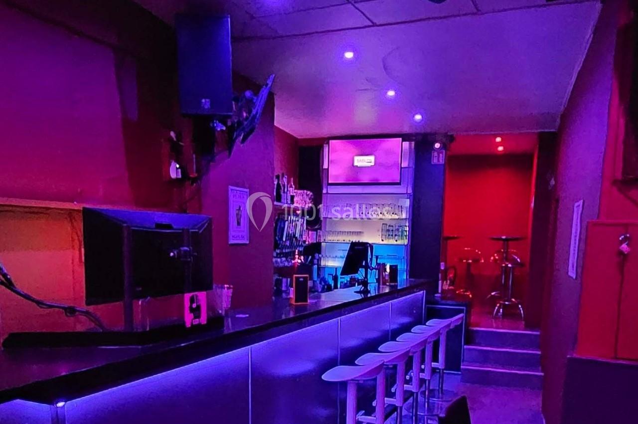 Intérieur d'un bar avec comptoir, tabourets blancs, éclairage violet et rouge, et étagères de bouteilles en arrière-plan.