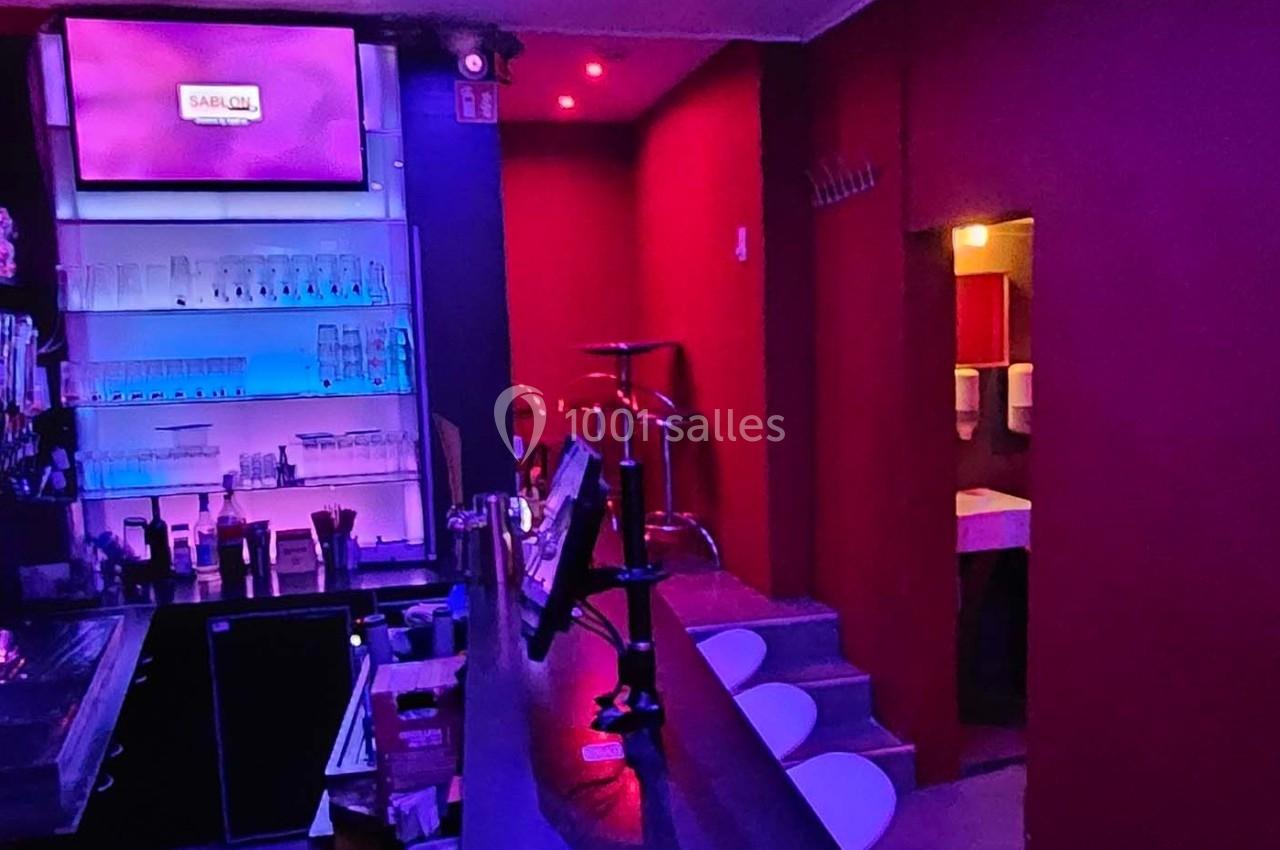 Intérieur d'un bar avec comptoir, étagères éclairées, tabourets et accès visible vers des toilettes.