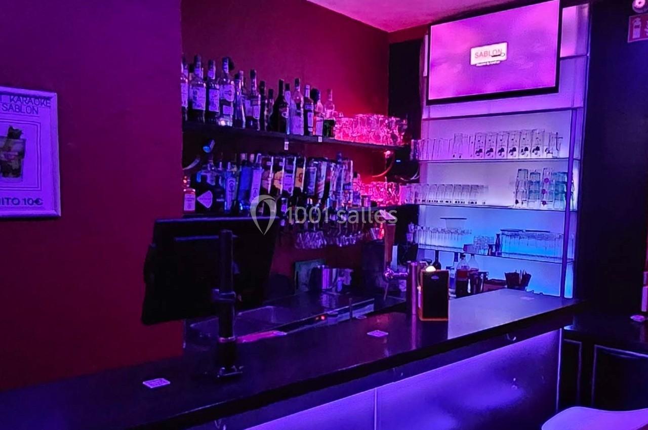 Bar avec comptoir éclairé, étagères remplies de bouteilles et verres, éclairage violet et écran mural allumé.