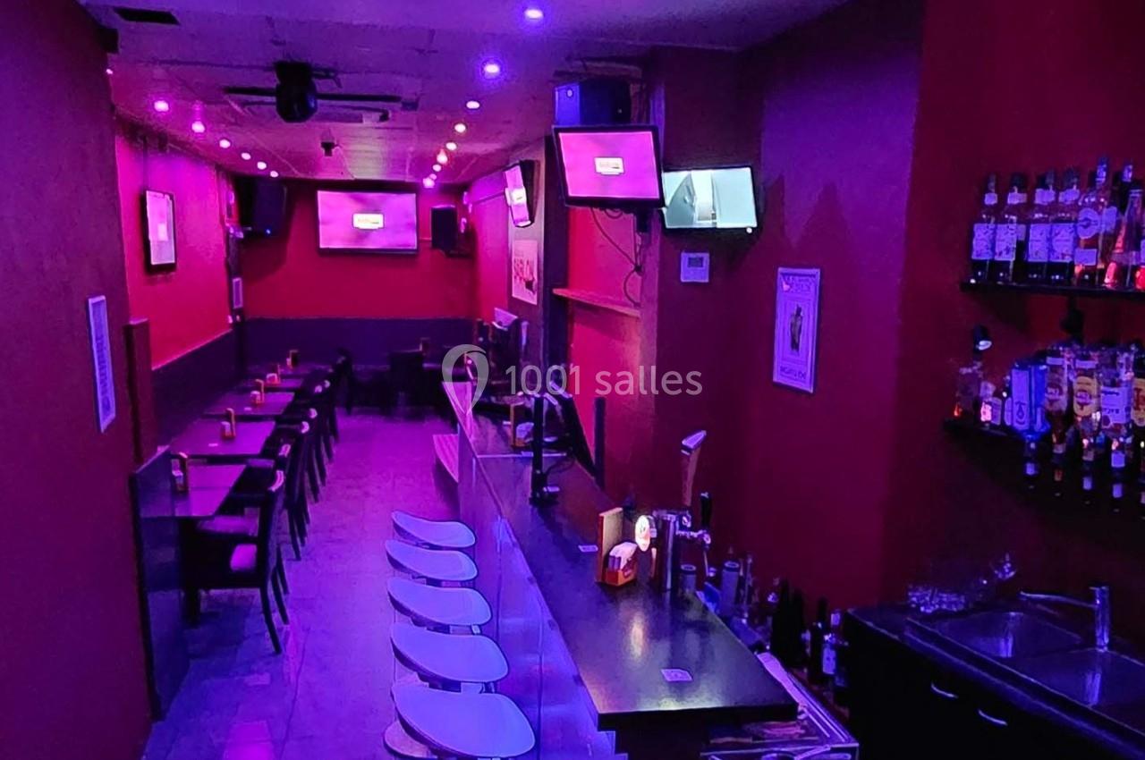 Bar avec éclairage violet, comptoir avec tabourets, étagères de bouteilles et tables alignées le long du mur.