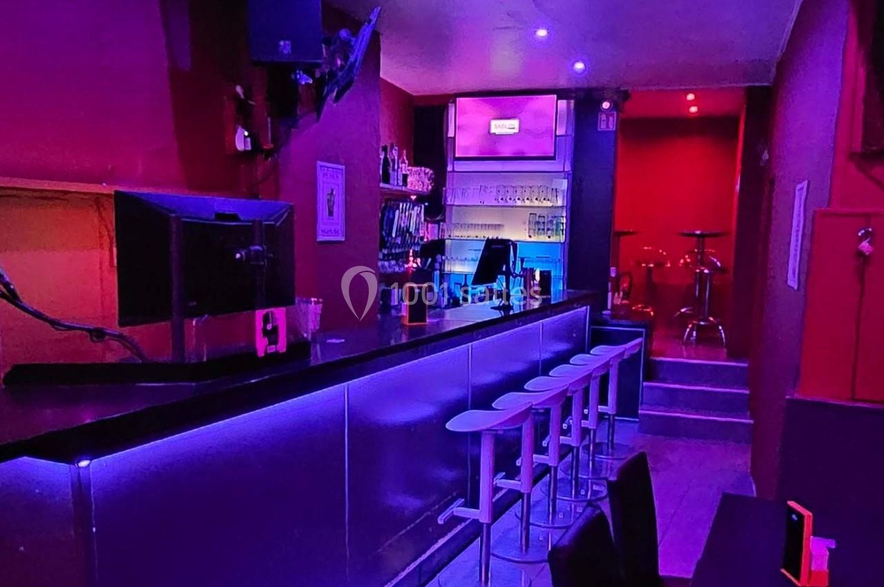 Bar avec comptoir éclairé en violet, tabourets alignés, étagères de boissons et ambiance lumineuse tamisée.