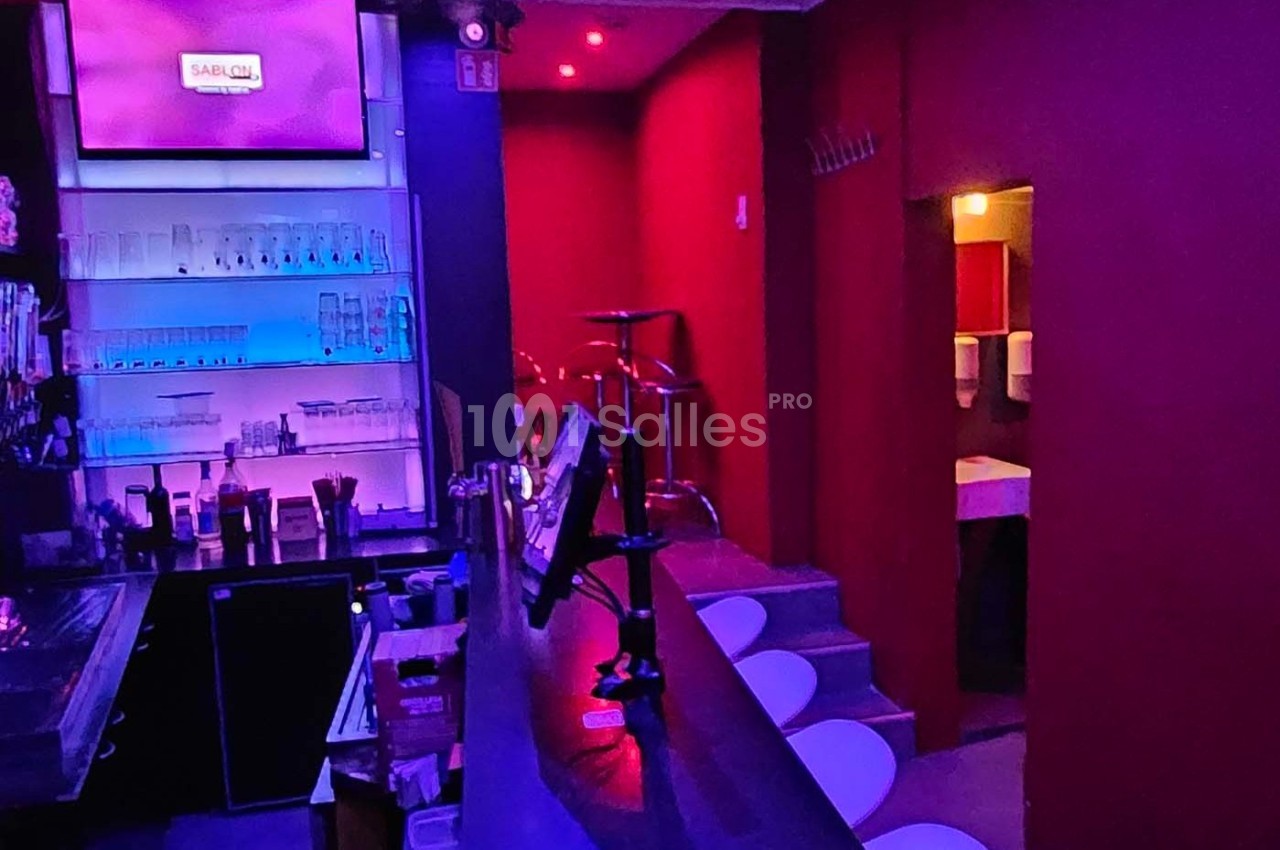Bar avec comptoir éclairé en bleu, tabourets blancs, étagères de verres et lumière tamisée rouge en arrière-plan.