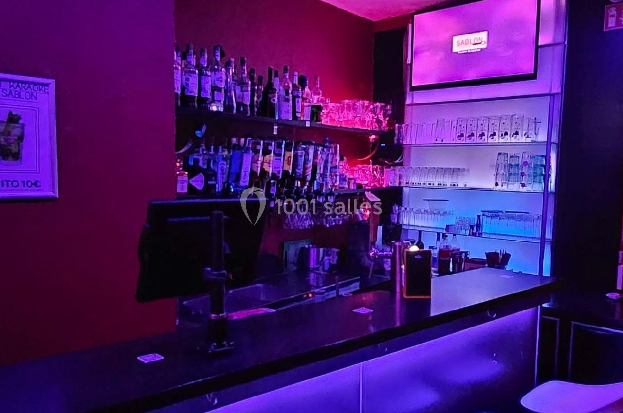 Bar avec comptoir éclairé, étagères remplies de bouteilles et verres, ambiance lumineuse violette et bleue.